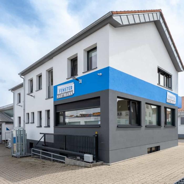 Fensterbau Hartmann Firmengebäude mit weißer Fassade, blauem Streifen und Logo, schwarze Fensterrahmen, und gepflasterter Vorplatz.