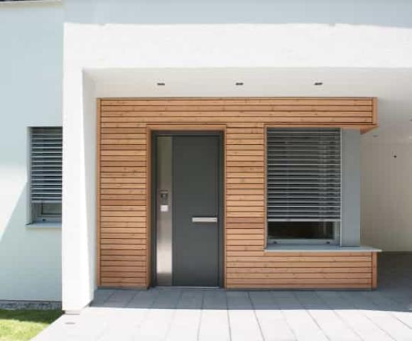 Moderner Hauseingang mit Holzverkleidung, dunkelgraue Haustür, großes Fenster mit Jalousien, und Pflastersteine vor grünem Rasen.