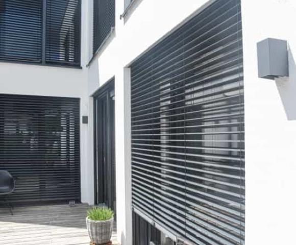 Terrassenbereich mit großen Fenstern und schwarzen Jalousien, Holzfußboden, moderner Stuhl, Pflanzgefäß, und weiße Fassade.