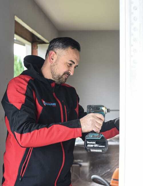 Fenstermonteur von Fensterbau Hartmann mit schwarzer und roter Arbeitsjacke, arbeitet mit Akkuschrauber am Fensterrahmen.